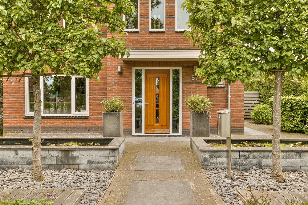 Front entrance of a brick house with a warm wooden door, flanked by two tall planters and green trees framing the doorway.