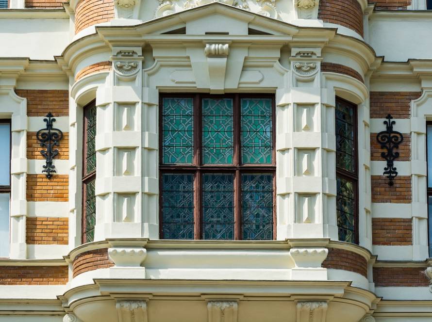 Curved bay window with lattice panes framed by cream stone on a brick building, with wrought-iron wall lamps on either side.