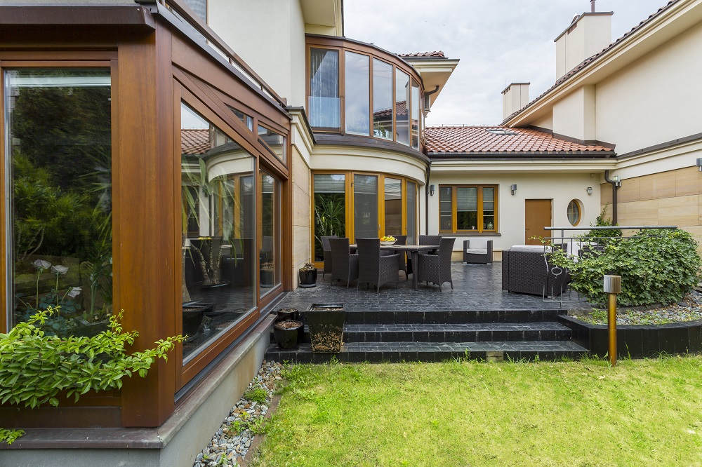 Backyard patio of a modern house with a round glass conservatory, outdoor dining table, and lounge area on a dark tiled deck.