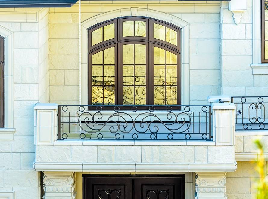 Stone house facade with a decorative wrought-iron balcony and large arched window above.
