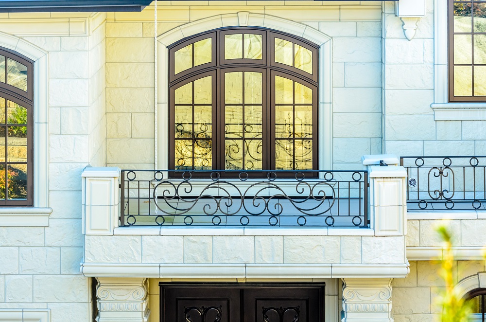 Stone house facade with a decorative wrought-iron balcony and large arched window above.