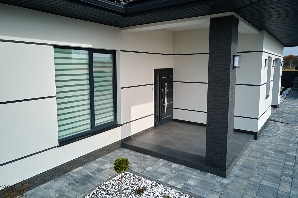 Modern house entrance with dark door under a covered porch, light walls with black trim, and a large window with frosted blinds to the left
