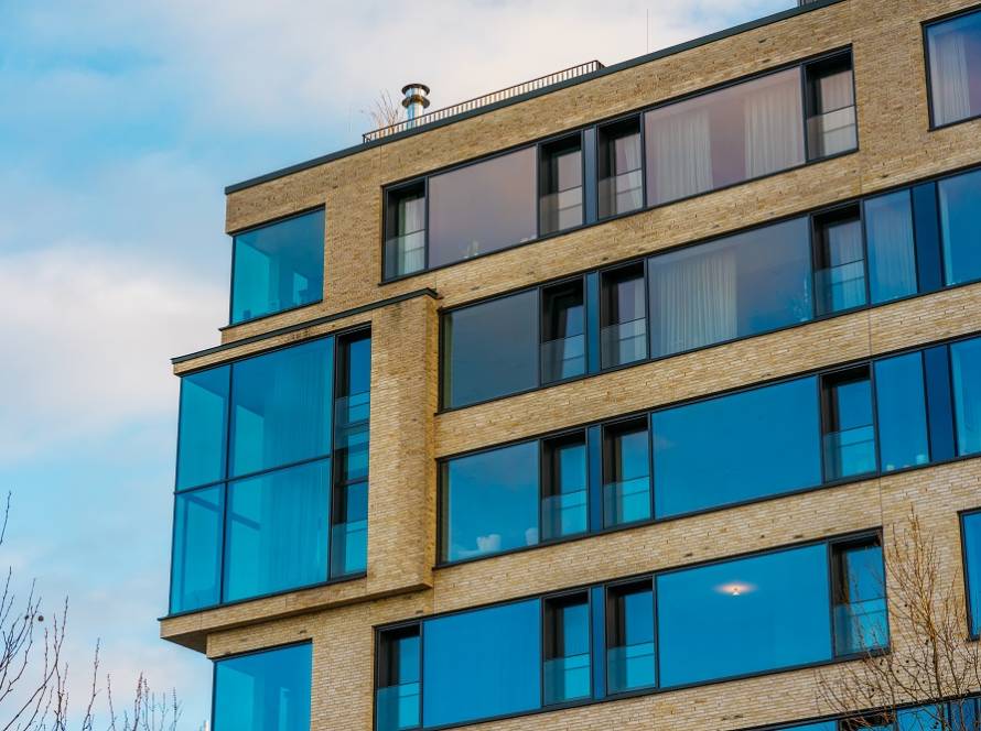 Exterior of a modern brick apartment building with large blue-tinted windows reflecting the sky and clouds, against a clear blue backdrop.