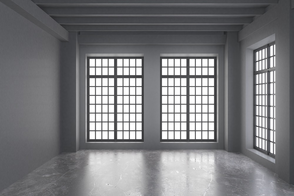 Empty industrial-style room with three tall grid windows letting in bright light, grey walls, and a reflective concrete floor.