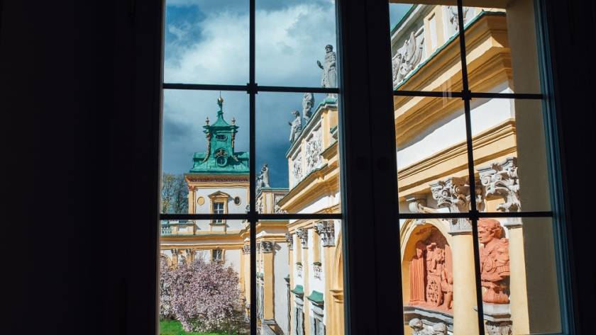 View through a window of an ornate yellow-and-white building with statues and a green-domed tower under a blue sky.
