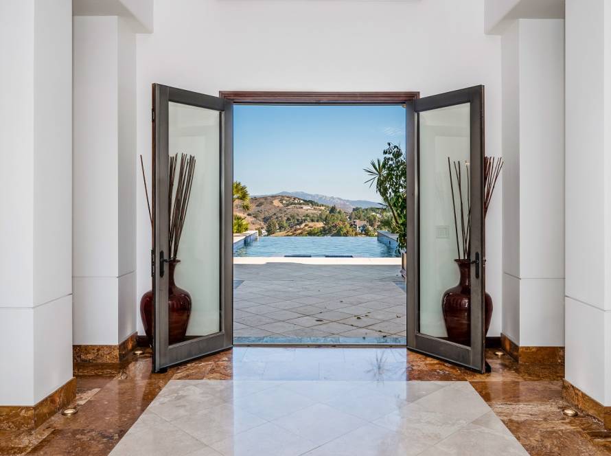 Open double doors reveal an infinity pool and hillside view, with marble floor and tall decorative vases inside moment.