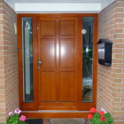 Wooden paneled front door with sidelights, brick entry, black wall mailbox, and pink and red flowers at the threshold.