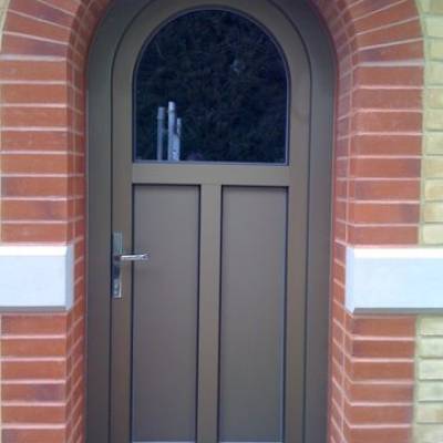Brown arched front door with a small upper curved window, set in a red brick arch on a yellow brick wall, with a metal door handle on the left.