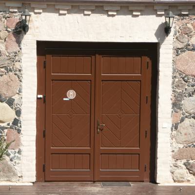 Double brown wooden doors with geometric panels set in a stone wall, flanked by two lanterns above the entrance.
