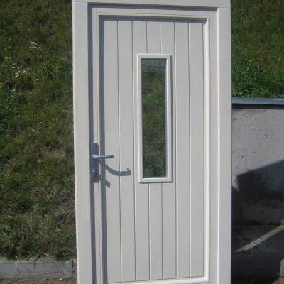 White exterior door with a narrow vertical glass pane in the center, silver handle, standing upright outdoors on pavement with grass behind.