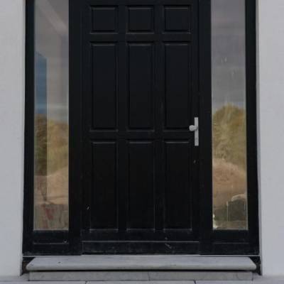 Black paneled front door with narrow glass sidelights, silver handle, set in a light wall with concrete steps and small blocks in front.