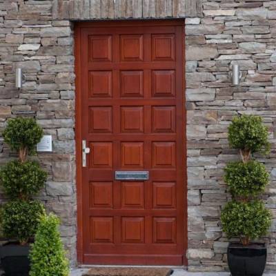 Red wooden front door with square panels set in a stone wall, flanked by trimmed topiary in pots.