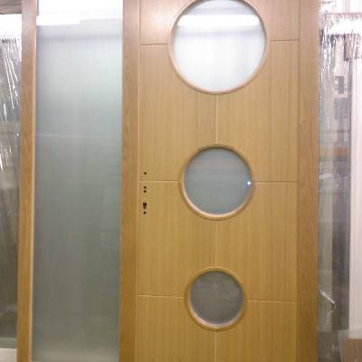 Wooden entry door with three circular glass windows stacked vertically and a frosted-glass panel on the left side.