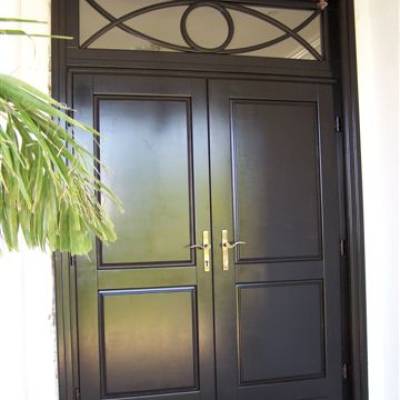 Double black front doors with brass handles, decorative transom panel above, a lantern light, and a white exterior with a leafy plant partially visible.