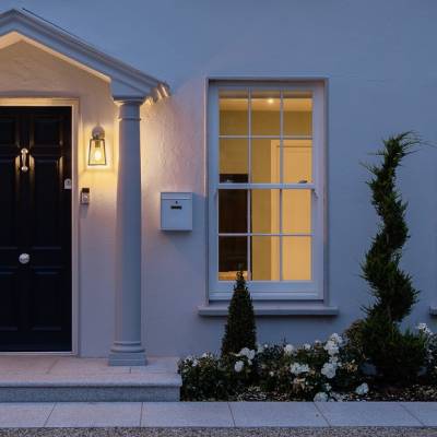 Front entrance of a blue house with a black door, two illuminated lanterns, and two columns framing the doorway, plus a small garden.
