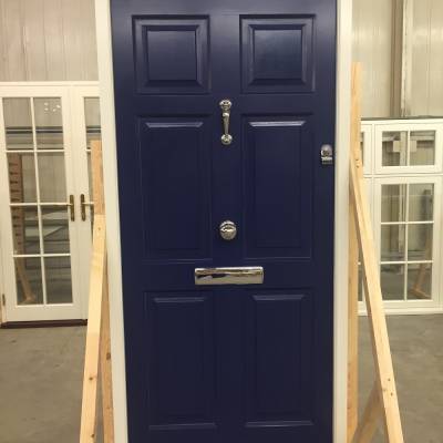 Blue six-panel front door with chrome handle, mail slot, and door knocker, mounted in a wooden display frame in a workshop showroom.